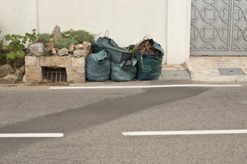 The image shows three large, dark green refuse bags filled with mixed waste, positioned on the edge of a paved street in an urban area, likely within Fitzrovia. The bags are situated next to a small garden bed with a cluster of plants and stones, which is bordered by a short brick or stone retaining wall. A grated drain is visible underneath the garden bed, indicating drainage for rainwater. The street surface appears clean and well-maintained, with white painted lines marking parking or lane boundaries. In the background, there is a white wall and part of a decorative metal gate or panel, hinting at residential or commercial premises. The scene is lit by natural daylight, with no visible shadows indicating overcast or diffused light conditions. The presence of the waste bags suggests a local rubbish collection or outdoor clearance, fitting within the context of services offered by Business Waste Removal Fitzrovia, serving the local area with rubbish removal solutions.