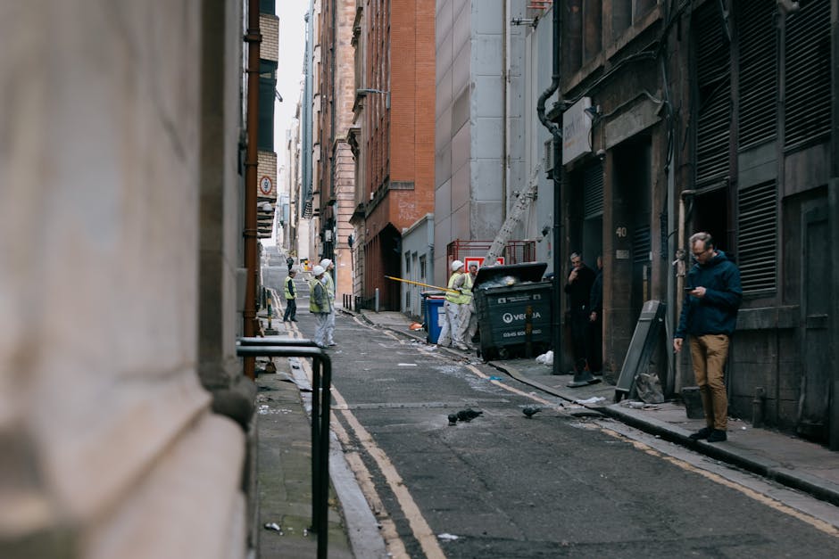 A large, used, beige cloth sack filled with various rubbish rests against the weathered exterior wall of an urban building on a narrow, uneven street. The bag appears to be made of thick, dirty fabric